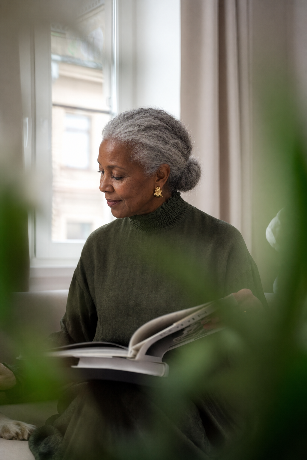 An older woman with silver curls reading a book beside a sunlit window, framed by green leaves
